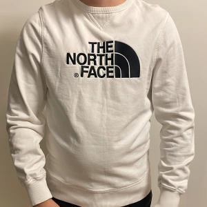 Vit sweatshirt från The North Face - Vit sweatshirt från The North Face med stor svart logga broderad på bröstet. Tröjan har rund hals, ribbade muddar och är långärmad. Materialet är mjuk bomull som känns skönt mot huden. Perfekt för chill dagar eller när du vill ha en clean streetstyle.