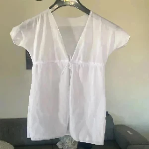 Vit kort omlott topp med snörning - Säljer en vit  topp i tunn och lätt bomull. Toppen har omlott-design med v-ringning och snörning framtill under bysten. Den är kortärmad och har en loose passform som ger en chill vibe. Perfekt att styla över linne. Supersnygg❤️