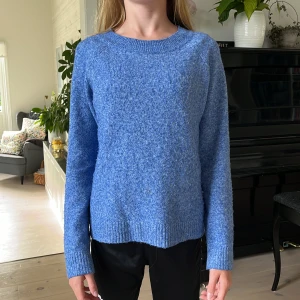 Blå stickad tröja med rund hals - Mysig blå stickad tröja med rundad hals och ribbade muddar vid ärmslut och nederkant. Tröjan har en lätt fluffig struktur och är långärmad, perfekt för kyliga dagar. Enkel och stilren design som är lätt att matcha med jeans eller mjukisbyxor.