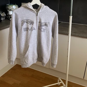 Grå hoodie med BVALDI-tryck - Snygg ljusgrå hoodie från BVALDI med coolt tryck av två händer och logga på bröstet. Tröjan har dragkedja, huva och två fickor framtill. Perfekt för chill dagar och streetstyle. Mjuk och skön i bomullsmix, långärmad och med relaxed passform. Ord-pris: 1099kr