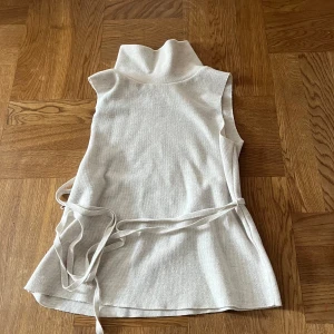 Beige stickad väst med knytband - Snygg beige stickad väst med hög krage och knytband i midjan. 