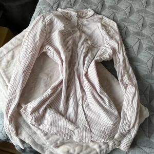 Randig skjorta från ICHI, stl 40 - Säljer en vit skjorta med tunna röda ränder från ICHI i storlek 40. Skjortan har klassisk krage, knäppning framtill och långa ärmar med manschett. Perfekt för en clean och stilren look. Materialet känns lätt och skönt mot huden.