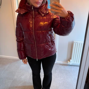 Glansig vinröd pufferjacka - Säljer en glansig vinröd pufferjacka med huva och dragkedja framtill. Jackan är vadderad och perfekt för kalla dagar. 