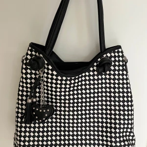 Svartvit axelväska med rutigt mönster - Snygg axelväska i svart och vitt med rutigt mönster och flätad look. Väskan har två rejäla handtag, dragkedja upptill och en cool tofs med hjärtformad detalj i svart med nitar. Perfekt för dig som gillar att sticka ut med accessoarer.