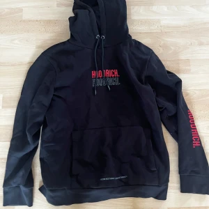 Svart hoodie från Hoodrich med tryck - Svart hoodie från Hoodrich med stor röd och grå logotyp på bröstet och ryggen. Snygga detaljer med snörning i huvan och tryck på ena ärmen. Praktisk ficka framtill och dragkedja på sidan av ärmen. Storlek L men passar M
