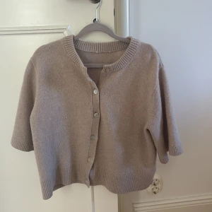 Beige kofta från & Other Stories - Superfin beige kofta från & Other Stories med korta ärmar och rund hals. Den har ribbade muddar och knappar framtill, perfekt att slänga på över en t-shirt eller linne. Mjuk och skön, stickad i ullblandning för extra komfort.