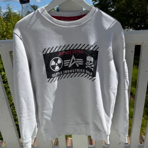 Vit sweatshirt från Alpha Industries - Säljer en vit sweatshirt från Alpha Industries med tryck på bröstet och ärmen. Framsidan har coolt grafiskt print med texten 'Do Not Enter' och logga, medan baksidan och ena ärmen har stora svarta bokstäver. Tröjan har rund hals och ribbade muddar.