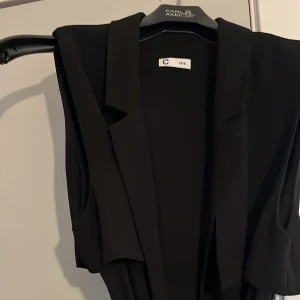 Svart lång väst med bälte från C - Säljer en svart lång väst från Cubs i storlek XS/S. Västen har en snygg krage, är ärmlös och kommer med ett matchande bälte i midjan. Perfekt att slänga över en skjorta eller t-shirt för en clean look.