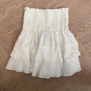 Vit volangkjol från Mayla Stockholm - Supersöt vit kjol från Mayla Stockholm med bred smock i midjan och dubbla volanger. Kjolen är lätt och luftig, perfekt för varma dagar. Super fint och lyxigt material köpt på nk förra året. Skriv gärna för fler bilder!