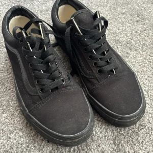 Svarta Vans sneakers med snörning - Säljer ett par klassiska svarta Vans sneakers med svart snörning och sula. Skorna har den ikoniska sidolinjen och är tillverkade i canvas och syntetmaterial. Perfekta för en stilren och avslappnad look.