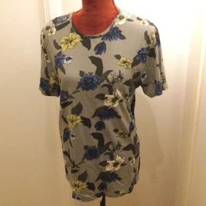 Blommig t-shirt i grått och blått M - Snygg t-shirt med blommigt mönster i blått, gult och vitt på grå botten. Klassisk rund halsringning och korta ärmar. Perfekt för en avslappnad och trendig stil. Blommig t shirt i bomull. Katter finns i hemmet!