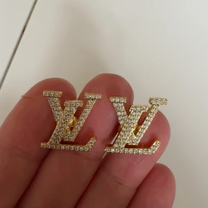 Luis vitton örhängen i guld med strass - Säljer ett par lyxiga LV örhängen i guldton med glittrande strass-stenar. Örhängena har den ikoniska LV-loggan och är perfekta för att ge din look extra glamour.