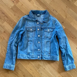 Blå jeansjacka från Lindex - Klassisk blå jeansjacka från Lindex i storlek 146. Jackan har knappar framtill, två bröstfickor med lock och knapp samt klassisk krage. 
