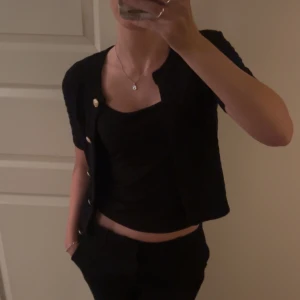 Svart stickad kofta - Kortärmad svart stickad kofta med ribbade kanter och dekorativa guldknappar framtill. Snygg och enkel att matcha till olika outfits. Perfekt för dig som gillar klassisk stil med en twist.