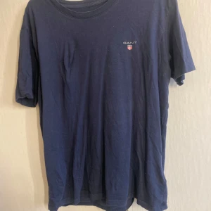 Mörkblå t-shirt från GANT - Mörkblå t-shirt från GANT med liten logga och sköld på bröstet. Klassisk rund halsringning och korta ärmar. Tillverkad i mjuk bomull, perfekt för dig som vill ha något snyggt men skönt. Även i jätte fint skick men den är skrynklig 🙈