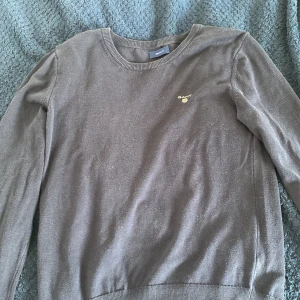 Mörkblå tröja från GANT - Säljer en klassisk grå tröja från GANT med rund halsringning och diskret logga broderad på bröstet. Tröjan har långa ärmar och ribbade muddar vid ärmslut och nederkant. Perfekt för en stilren vardagslook.