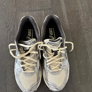 Gråa Asics sneakers med mesh och snörning - Säljer ett par gråa Asics sneakers med vita och svarta detaljer. Skorna har meshpaneler för extra ventilation och klassisk snörning. Perfekta för träning eller vardagsstil. Snygg sportig design med bekväm passform.