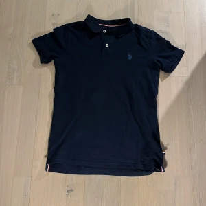 Mörkblå pikétröja från US Polo Assn - Säljer en klassisk mörkblå pikétröja från US Polo Assn med broderad logga på bröstet. Tröjan har korta ärmar, krage och knäppning framtill. Detaljer i rött och vitt på insidan av kragen ger en sportig touch.