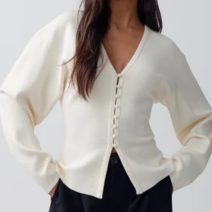 Vit kofta med knappar - Säljer en stilren vit kofta med v-ringning och dekorativa knappar framtill. Tröjan har lång ärm och är tillverkad i ett mjukt stickat material. Jag beställde den men aldrig använt. Prislappen är kvar på tröjan!