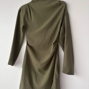 Olivgrön långärmad klänning med drapering - Säljer en olivgrön klänning med lång ärm och snygg drapering i midjan. Klänningen har en enkel, stilren design och är tillverkad i ett mjukt material som ger en bekväm passform. Perfekt för dig som gillar minimalistisk stil.