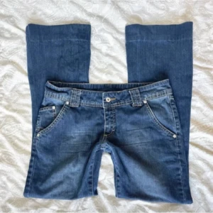 Blå lågmidjade bootcut jeans  - Säljer ett par blå lågmidjade bootcut jeans med fem fickor och snygga detaljer på ena bakfickan. Jeansen har normal passform och är tillverkade i denim med lätt tvättade detaljer. Perfekta för dig som gillar en tidlös stil. Midjemåttet är 41cm och innerbenslängden 75cm men de går att ta ner minst 5 cm!