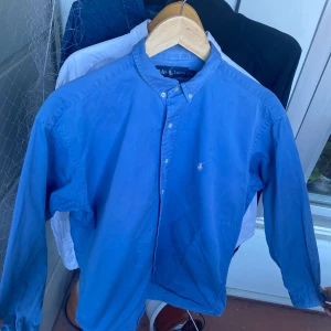 Blå skjorta från ralph Lauren - Säljer en klassisk blå skjorta från Ralph Lauren i modellen Blake. Skjortan har lång ärm, button-down krage och den ikoniska broderade loggan på bröstet. Tillverkad i 100% bomull för en skön och stilren känsla. Storlek S