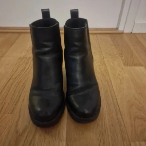 Svarta boots med klack - Säljer ett par svarta boots i skinn med rund tå och klack. Skorna har en enkel och stilren design med dragflikar upptill för enkel påtagning. Perfekta för dig som vill ha ett par klassiska boots till garderoben.