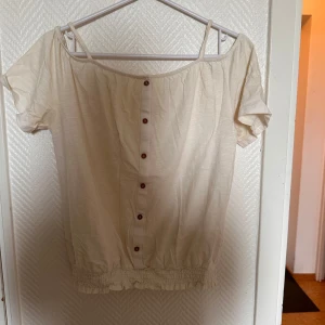 Offshoulder topp med knappar från H&M - Säljer en ljusbeige offshoulder topp från H&M med dekorativa knappar framtill. Toppen har smala axelband och resår i nederkant för en snygg passform. Perfekt för varma dagar!