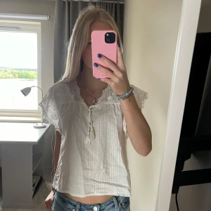 Vit blus med spetsdetaljer - Säljer en söt vit blus med spets över axlarna och volangkant. Blusen har korta ärmar och knytning framtill vid halsen. 