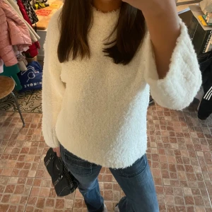 Vit fluffig stickad tröja - Mysig vit stickad tröja med fluffig textur och lång ärm. Perfekt för kyligare dagar och enkel att matcha med jeans eller kjol. Tröjan har en rak passform och rund halsringning.