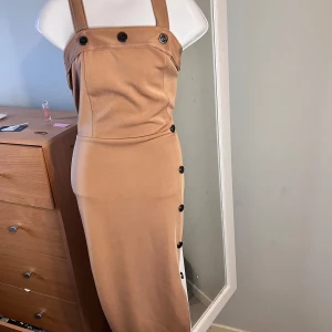 Beige  body klänning med knappar - Snygg beige klänning med knappar framtill och smala axelband. Klänningen har en slits och en figurnära passform som framhäver silhuetten. Perfekt för en stilren look.