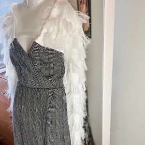 Mönstrad jumpsuit med fjäderdetaljer - Snygg jumpsuit med svartvitt mönster och unika vita fjäderärmar. Den har en omlott-design och v-ringning som ger en elegant touch. Perfekt för att sticka ut på festen!                en liten fläck på klänning ( bild)