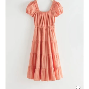 Persikofärgad klänning med puffärm - Söt persikofärgad klänning med kort puffärm, fyrkantig ringning och volangdetaljer. Klänningen har öppen rygg med knytband och är tillverkad i ett lätt, luftigt material. Perfekt för varma dagar och sommarfester. Endast använd 2 gånger. Nypris 1450 kr