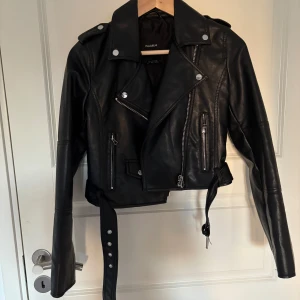 Svart skinnjacka från Pull&Bear - Säljer en svart skinnjacka från Pull&Bear. Jackan har dragkedjor, knappar, axelklaffar och bälte i midjan🌸