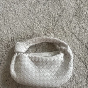 Vit flätad handväska med knutdetalj - Säljer en snygg vit handväska med flätat mönster och en unik knutdetalj på handtaget. Väskan har dragkedja upptill och är perfekt för att bära det viktigaste. Stilren och trendig accessoar som lyfter varje outfit.