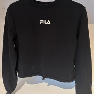 Svart croppad sweatshirt från FILA - Svart sweatshirt från FILA med vit logga framtill och färgglad logga på ryggen. Tröjan är croppad med dragsko nertill och har långa ärmar. Perfekt för en sportig och avslappnad stil.