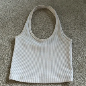 Vit ribbad halterneck linnetopp från H&M Divided - Säljer en vit ribbad halterneck linnetopp från H&M Divided. Toppen har en croppad passform och är ärmlös med breda axelband som går runt nacken. Perfekt för varma dagar eller som layering-plagg.