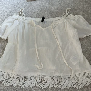 Vit blus med spetsdetaljer från H&M - Säljer en söt vit blus från H&M med tunna axelband och offshoulder-design. Blusen har knytband framtill och vacker spetskant nertill med blommönster. Perfekt för varma dagar och somriga outfits!