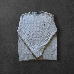 Ljusgrå kabelstickad tröja från Ralph Lauren - Kabelstickad ljusgrå tröja från Ralph Lauren med rund halsringning och broderad logga på bröstet. Tröjan har ribbade muddar vid ärmslut och nederkant. Perfekt för lager-på-lager och klassisk stil. Den är i storlek S men passar även Xs