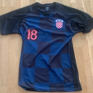 Kroatiens fotbollströja  - Säljer en officiell Kroatien fotbollströja med Rebic och nummer 18 på ryggen. Tröjan är blå med mörkblå rutmönster, kortärmad och har Kroatiens landslagsemblem på bröstet. Perfekt för fotbollsfans!