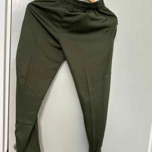 Mörkgröna träningsbyxor från Jordan x PSG - Snygga mörkgröna träningsbyxor med elastisk midja och tryckta loggor från Paris Saint-Germain och Jordan. Byxorna har smal passform och detaljer vid knäna. Perfekta för sport eller chill.