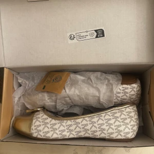 Michael Kors ballerinaskor med rosett och hjärtdetalj - Säljer ett par ballerinaskor från Michael Kors i färgen vanilj/guld. Skorna har ett diskret MK-mönster, guldfärgad tå, rosett framtill och en liten hjärtdetalj. Bekväm quiltad innersula och platt sula. Perfekta för dig som gillar stilrena detaljer.