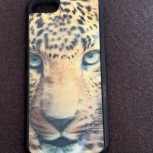 iPhone 7 mobilskal  - Det här är ett mobilskal med ett realistiskt tryck av ett leopardansikte. Skalet är svart runt kanterna och har ett färgstarkt motiv som passar dig som vill sticka ut och visa din vilda sida. Perfekt för dig som gillar djur och vill ge din mobil en unik look. Tror den är till iPhone 7. Slitet mobilskal som börjat gå upp se bild