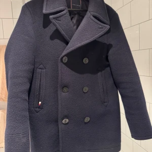 Mörkblå skepparkavaj från Tommy Hilfiger - Stilren mörkblå (100% ull) skepparkavaj från Tommy Hilfiger med dubbelknäppning och bred krage. Jackan har två snedställda fickor framtill och klassisk marin känsla. Perfekt för kyligare dagar och ger en tidlös look. Vill minnas att nypriset var ca 4500, storlek S passar M. Utmärkt skick, inget som tyder på användning. 185 lång 75 kg