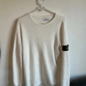 Vit stickad tröja från Stone Island - Vit stickad tröja från Stone Island med klassisk svart logotyp-patch på vänster ärm. Tröjan har rund halsringning och ribbade muddar. Perfekt för dig som gillar stilrena och exklusiva plagg.
