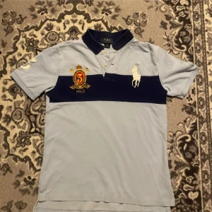  pikétröja från Polo Ralph Lauren - Säljer en ljusblå pikétröja från Polo Ralph Lauren med mörkblå krage och bred rand över bröstet. Tröjan har broderad logga och emblem med siffran 3 på bröstet. Klassisk piké med korta ärmar och knappar vid halsen. Bara att höra av sig vid funderingar!