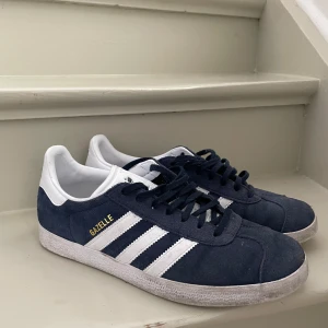 Adidas Gazelle mörkblå sneakers - Säljer ett par klassiska Adidas Gazelle sneakers i mörkblå mocka med vita detaljer och de ikoniska tre ränderna på sidorna. Skorna har snörning och vit platt sula. Perfekta för en stilren och sportig look.