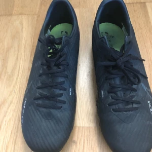 Svarta Nike Air Zoom fotbollsskor - Säljer ett par svarta Nike Air Zoom fotbollsskor med snörning och rund tå. Skorna har en låg profil och är designade för bra grepp på planen. Snygg och sportig look med diskreta detaljer och grönt foder på insidan.