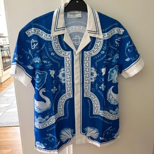 Helt ny skjorta från Casablanca - Säljer en helt ny blå och vit skjorta från Casablanca med korta ärmar och klassisk krage. Skjortan har ett detaljerat mönster med svanar, blommor och dekorativa ramar. Perfekt för dig som vill sticka ut med en unik och färgstark look.