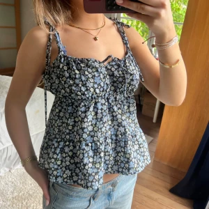 Blommig linnetopp med knytband - Säljer en söt blommig linnetopp med blå, vita och gula blommor. Toppen har smala axelband med knyt, v-ringning och en liten rosett framtill. Passformen är lös och luftig, perfekt för varma dagar.